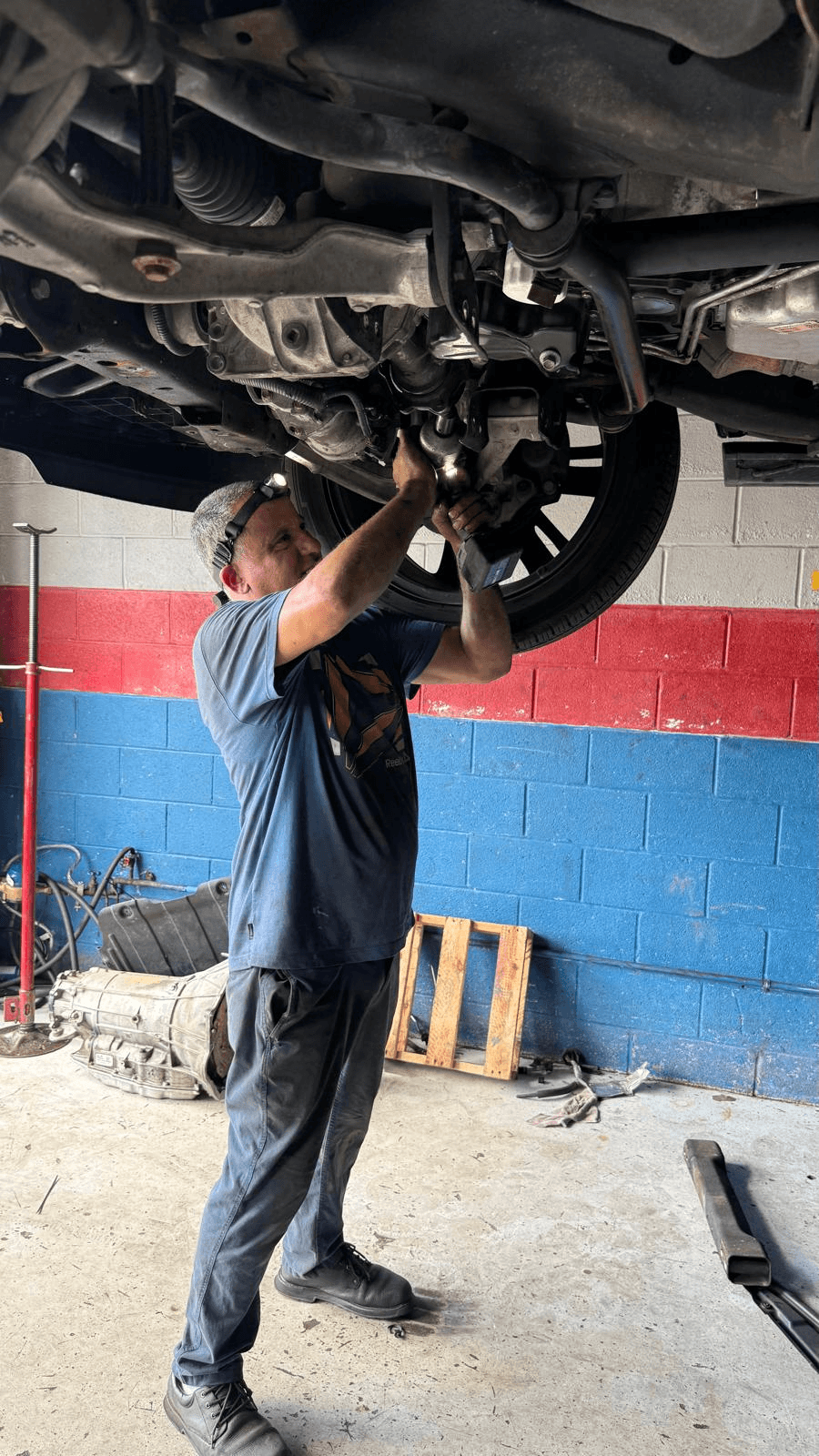 A technician working underneath a raised vehicle at Instant Auto Repair, with the exhaust and driveline visible and a transmission on the shop floor